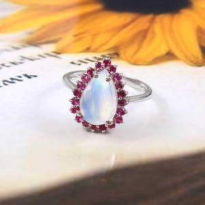 May include: A pearlescent moonstone ring with a teardrop-shaped stone surrounded by a halo of small red rubies.