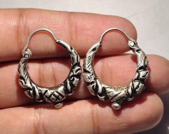 May include: A pair of silver-toned hoop earrings. Each earring features an ornate, textured design with floral and geometric patterns. The earrings have a simple hook closure and a small, rounded pendant at the bottom. The earrings are displayed against a neutral background.