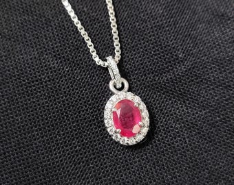 May include: A silver necklace with a pendant featuring a large oval ruby surrounded by small, sparkling diamonds. The ruby is a deep red colour, and the diamonds are clear. The chain is a delicate silver link.