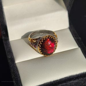 May include: A gold and silver ring featuring a large, oval, deep red gemstone. The ring has detailed patterns around the gemstone and on the band. It is displayed in a black jewellery box with a white interior.