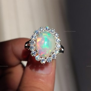 Puede incluir: Un anillo de plata con una gran piedra de ópalo ovalada con colores iridiscentes. El ópalo está rodeado por un halo de pequeñas piedras preciosas transparentes. La banda del anillo tiene un diseño simple de plata pulida. Es una joya.