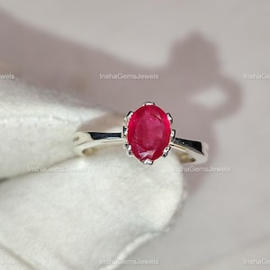 Könnte beinhalten: Ein silberner Ring mit einem leuchtenden, oval geschliffenen Rubin. Der Rubin ist in einer Krappenfassung gehalten, die seine tiefrote Farbe zur Geltung bringt. Der Ring hat ein schlichtes, poliertes Silberband.