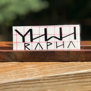 May include: A rectangular dark brown wooden stand holding a white decal with a red grid pattern. The decal features black stylized lettering that reads YHWH above the word RAPHA in a geometric font style with sharp, angular lines and characters.