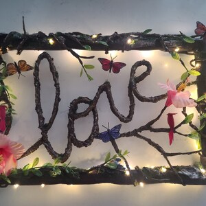 May include: A wooden frame with the word "love" spelled out in twigs, decorated with butterflies, flowers, and fairy lights.