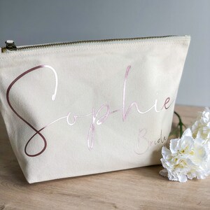 May include: A white canvas makeup bag with a zipper closure. The bag has the name "Sophie" written in pink script with the word "Bride" underneath.