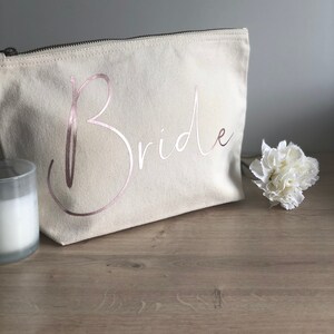 May include: A cream canvas makeup bag with a zip. The word "Bride" is printed in rose gold script. A white flower and a candle in a glass holder are next to the bag. The bag is on a wooden surface.