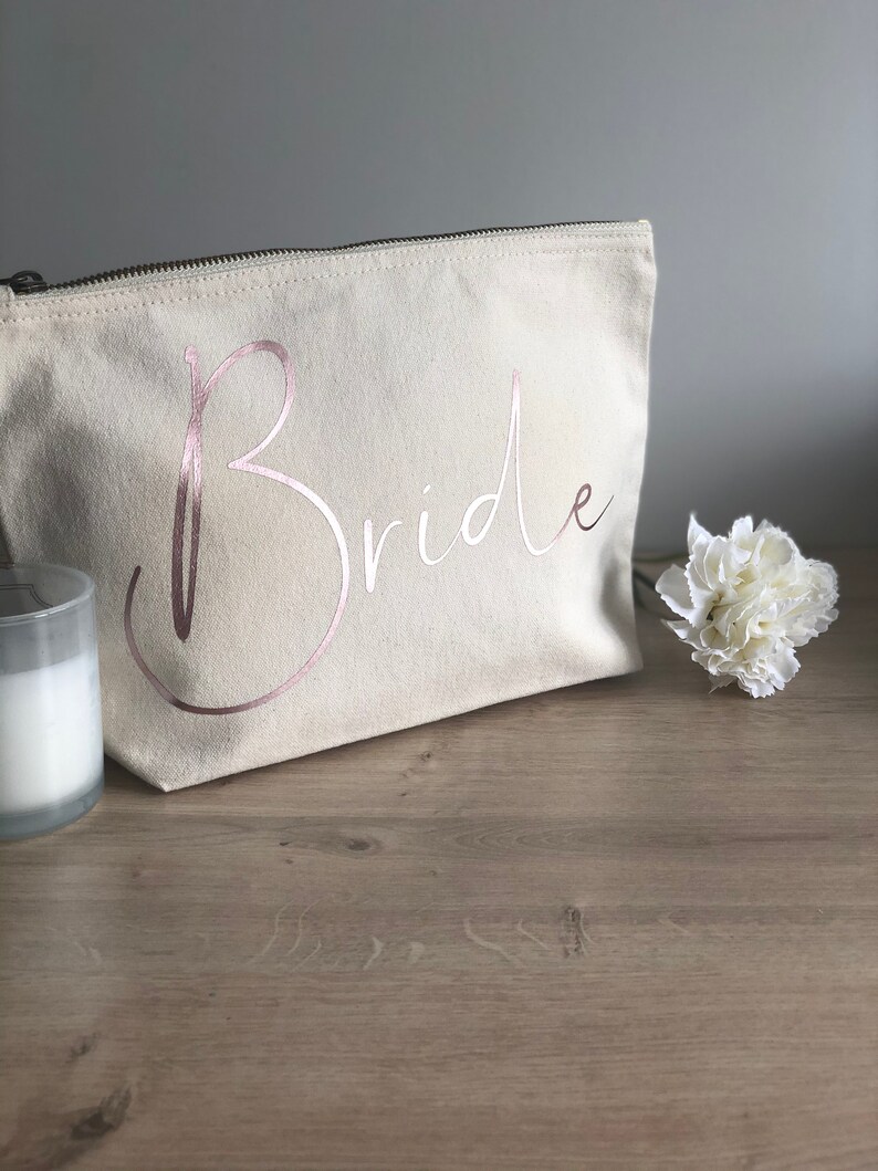 May include: A cream canvas makeup bag with a zip. The word "Bride" is printed in rose gold script. A white flower and a candle in a glass holder are next to the bag. The bag is on a wooden surface.