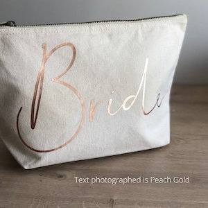 May include: A cream canvas makeup bag with a black zip. The word "Bride" is written in rose gold script across the front. The text "Text photographed is Peach Gold" is below the bag.