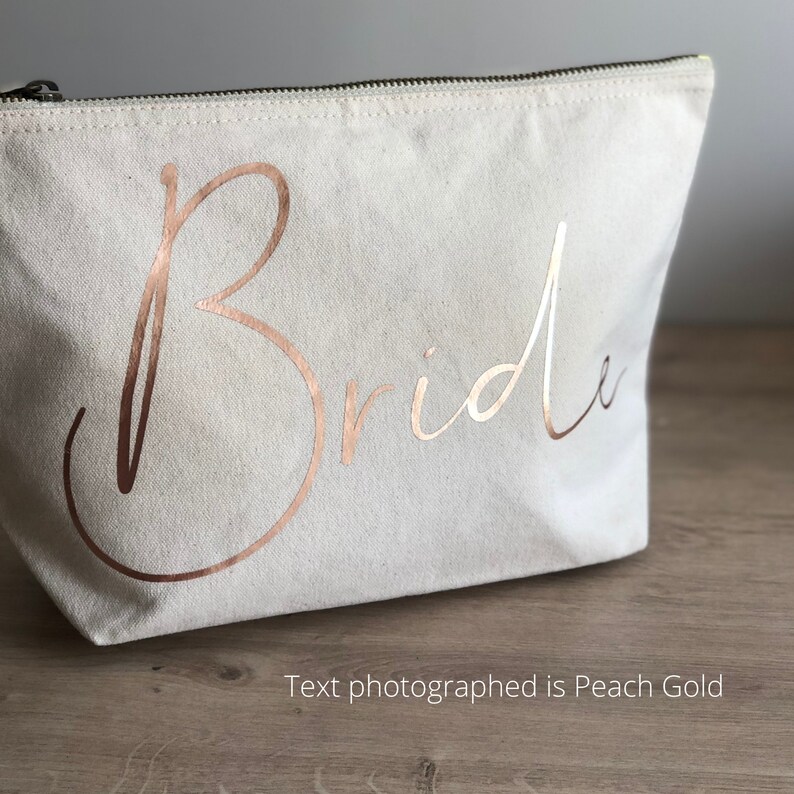 May include: A cream canvas makeup bag with a black zip. The word "Bride" is written in rose gold script across the front. The text "Text photographed is Peach Gold" is below the bag.