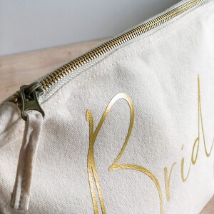 May include: A cream-coloured canvas cosmetic bag with a gold zip and the word "Bride" in gold script. The bag is rectangular and has a tassel attached to the zip pull.