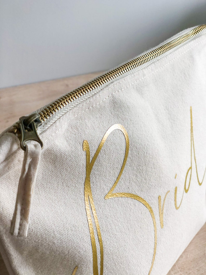 May include: A cream-coloured canvas cosmetic bag with a gold zip and the word "Bride" in gold script. The bag is rectangular and has a tassel attached to the zip pull.