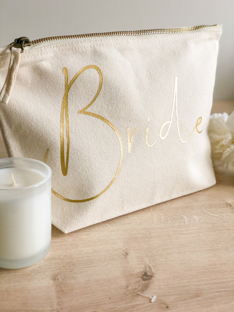 May include: A cream-coloured canvas cosmetic bag with a gold zip and the word "Bride" in gold script. A clear glass candle holder with a white candle is to the left of the bag. The bag is on a wooden surface.