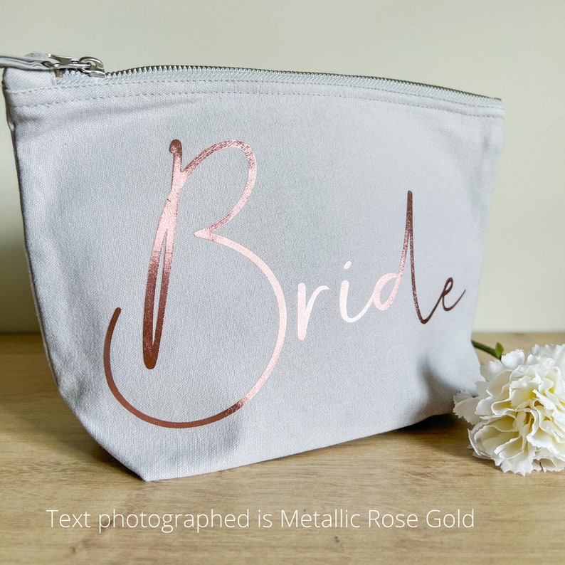 May include: A pale grey canvas makeup bag with a zip closure. The word "Bride" is printed in metallic rose gold script across the front. A white flower rests on the wooden surface next to the bag. The text photographed is Metallic Rose Gold.
