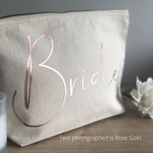 May include: A beige canvas makeup bag with the word "Bride" in a rose gold script. The bag has a zip closure. The text photographed is Rose Gold. The bag is on a wooden surface.