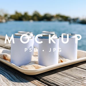 May include: Three blank white foam can coolers sit upright on a light-colored fabric cloth inside a rectangular tray. The text MOCKUP PSD . JPG is overlaid in white across the center of the image, positioned over the can coolers.