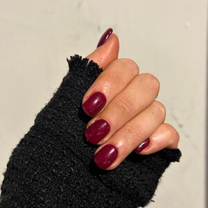 Peut inclure: Gros plan sur une main avec des ongles courts et arrondis peints d'un rouge bordeaux foncé.