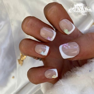 May include: Close-up of a hand with short, square-shaped nails painted with a nude polish and a white french tip. Each nail has a small, pastel-coloured flower design on the tip.