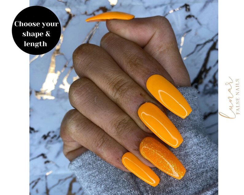 May include: A hand model wearing long, bright orange press-on nails with a glittery accent nail. The nails are long and almond shaped. The text "Choose your shape & length" is visible in the image.