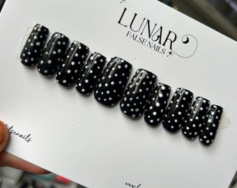 May include: A set of ten black press-on nails with white polka dots. The nails are arranged in a row on a white card with the text "LUNAR FALSE NAILS" and a website address.
