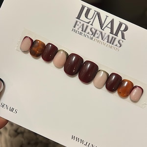 May include: A set of ten false nails in a burgundy, orange, and nude colour scheme. The nails have a gold accent and are displayed on a white card with the text "LUNAR FALSENAILS PREMIUM NAIL ENHANCEMENTS" and "WWW.LUNARFALSENAILS.COM".