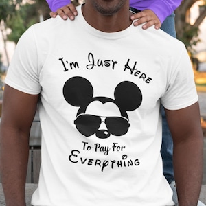May include: A white t-shirt featuring a black Mickey Mouse wearing sunglasses and the text "I'm Just Here To Pay For Everything".