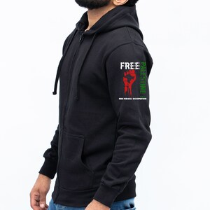 Pode incluir: Casaco de moletom preto com z&iacute;per e capuz, um punho vermelho e as palavras "Free Palestine" e "End Israeli Occupation" impressas na manga.