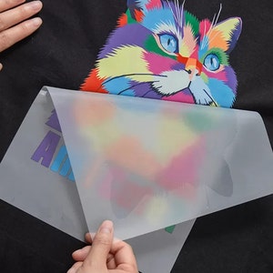 May include: A clear plastic sheet is being peeled off of a black t-shirt with a colorful cat graphic. The cat is a cartoon style with a rainbow of colors. The t-shirt has the word "Animal" printed on it.