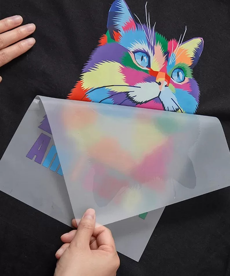 May include: A clear plastic sheet is being peeled off of a black t-shirt with a colorful cat graphic. The cat is a cartoon style with a rainbow of colors. The t-shirt has the word "Animal" printed on it.