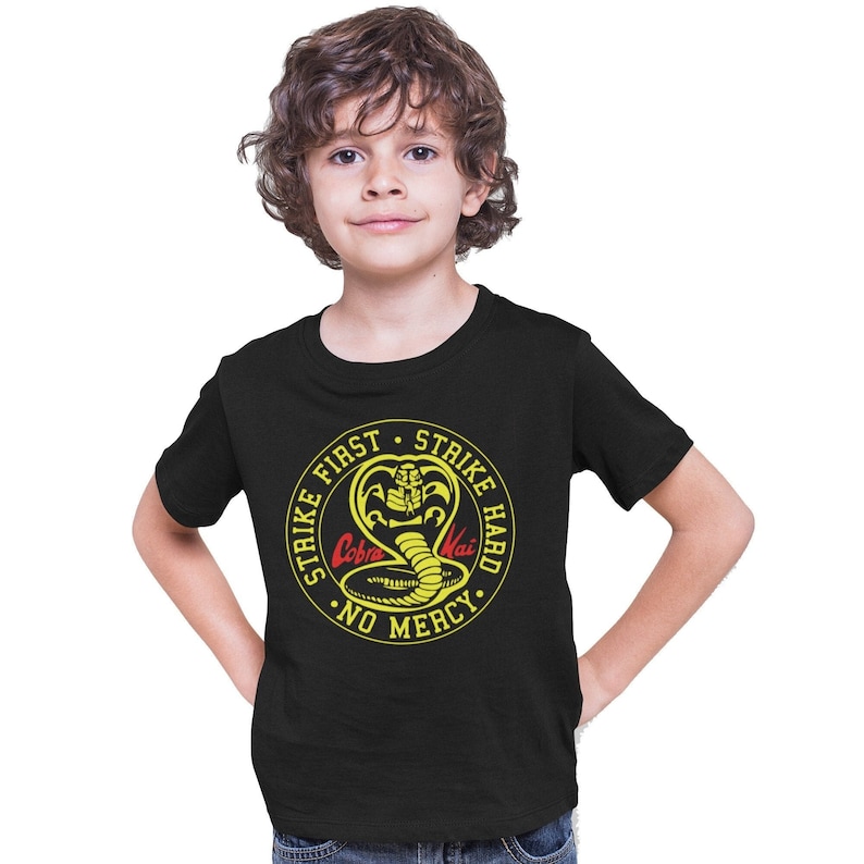 May include: Black t-shirt with a yellow circle graphic featuring a cobra with the text "Strike First Strike Hard No Mercy" and "Cobra Kai" in red.