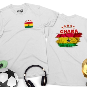 May include: Two white t-shirts with a Ghanaian flag design. One shirt has a small flag on the front, the other has a larger flag with the word "GHANA" on the back. The shirts are laid out on a white surface with a soccer ball, headphones, and a gold soccer trophy.