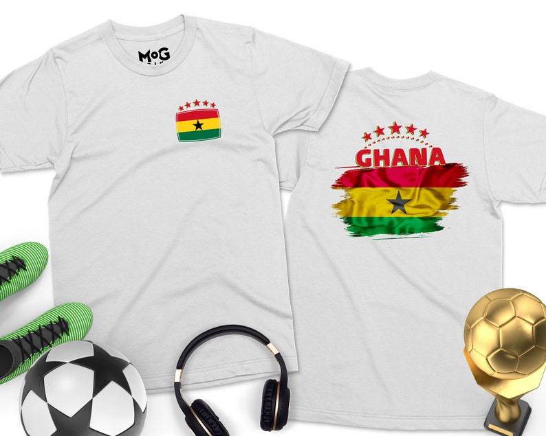 May include: Two white t-shirts with a Ghanaian flag design. One shirt has a small flag on the front, the other has a larger flag with the word "GHANA" on the back. The shirts are laid out on a white surface with a soccer ball, headphones, and a gold soccer trophy.