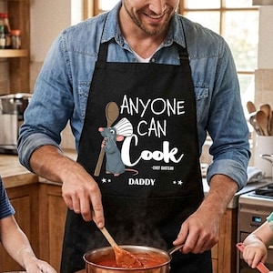 Könnte beinhalten: Schwarze Schürze mit dem Text "ANYONE CAN COOK - Chef Gusteau DADDY" und einer Cartoon-Ratte, die einen Kochlöffel hält. Eine Person trägt die Schürze beim Kochen in einer Küche.