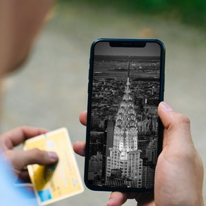 Puede incluir: Una persona sosteniendo un teléfono inteligente que muestra una foto en blanco y negro del edificio Chrysler en la ciudad de Nueva York. La persona también sostiene una tarjeta de crédito amarilla.