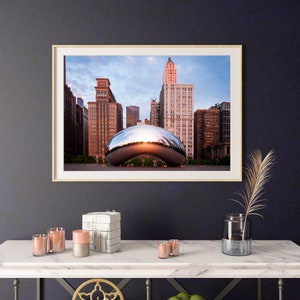 May include: A framed photograph of the Cloud Gate sculpture in Chicago, also known as "The Bean", reflecting the city skyline at sunset. The sculpture is a large, shiny, bean-shaped structure made of stainless steel.