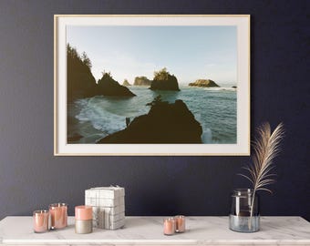 Op de afbeelding: Ingelijste landschapsafdruk met een kustlandschap met donkere rotsformaties, oceaangolven en een lichtblauwe lucht. De afdruk staat in een gouden lijst en hangt boven een tafel met marmeren blad met kaarsen en decoratieve items.