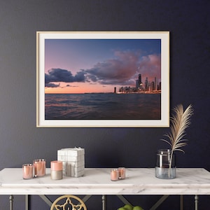 May include: A framed photograph of a city skyline at sunset. The city is on the shore of a body of water. The sky is a mix of pink, purple, and blue with fluffy clouds. The city lights are reflected in the water.
