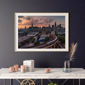 Può includere: Una vista aerea di uno skyline cittadino con un treno che viaggia su un binario curvo in primo piano. Il cielo è di un rosa e un arancio vibranti, con nuvole che riflettono il tramonto.