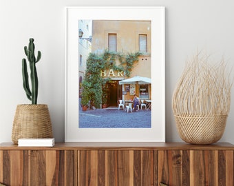 May include: A framed photograph of a small cafe in Italy with a sign that reads "BAR" above the entrance. The cafe is surrounded by greenery and has a few tables and chairs set up outside. A person is sitting at one of the tables.
