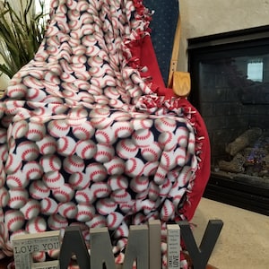 May include: A baseball-themed fleece blanket draped over a blue armchair. The blanket features a repeating pattern of baseballs with red stitching on a white background. The blanket is trimmed with red fringe. Wooden letters spell "FAMILY" in front of the chair.