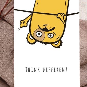 May include: A white card with the text "THINK DIFFERENT" features a yellow cartoon character hanging upside down from a black line. The character has a surprised expression. Dried flowers and a brown cloth are in the background.