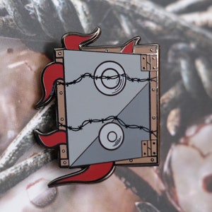 Könnte beinhalten: Quadratischer Emaille-Pin mit grauem und beigem Design. Der Pin zeigt zwei weiße Kreise mit einem schwarzen Stacheldraht-Design und roten flammenartigen Akzenten. Der Pin hat einen silbernen Rand.
