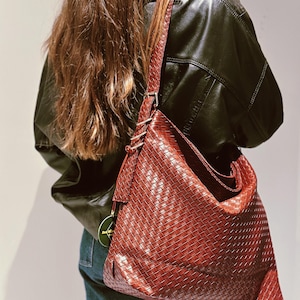 Può includere: Una borsa a tracolla intrecciata marrone rossiccio con una lunga tracolla. La borsa ha un design ampio e morbido e si indossa a spalla. La borsa è realizzata in materiale intrecciato e la tracolla è regolabile. La borsa è abbinata a una giacca di pelle nera e jeans.