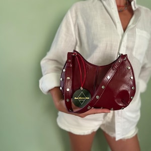 Peut inclure: Un sac à bandoulière bordeaux avec des clous argentés sur les bords. Le sac a une forme incurvée et une sangle assortie. Une étiquette ronde avec du texte est suspendue à la sangle. Le sac est tenu contre une chemise blanche.