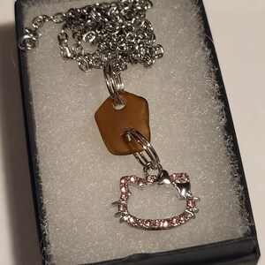 May include: A silver chain necklace with a brown sea glass charm and a silver charm shaped like Hello Kitty with pink rhinestones.