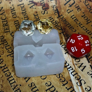 May include: Two clear resin dice with a yellow and a clear color, sitting in a white silicone mold. A red twenty-sided die with black numbers is on a brown surface.