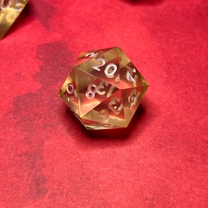 May include: A clear, yellow, 20-sided dice with white numbers on each side. The dice is sitting on a red surface.