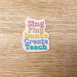 May include: A pink sticker with the words "Sing, Play, Dance, Create, Teach" in colorful, retro-style lettering. The sticker has a rounded, cloud-like shape and is set against a light wood grain background.