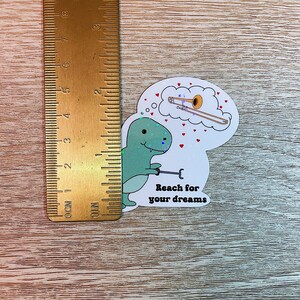 May include: A green dinosaur sticker with a thought bubble above its head. The thought bubble contains a gold trombone. The sticker says "Reach for your dreams".