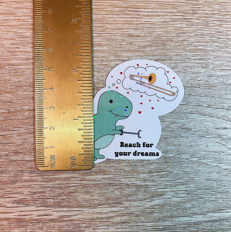 May include: A green dinosaur sticker with a thought bubble above its head. The thought bubble contains a gold trombone. The sticker says "Reach for your dreams".