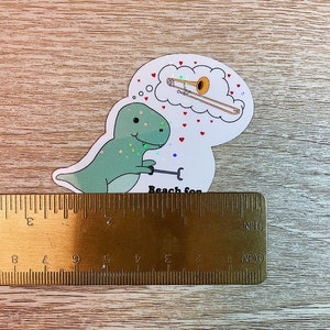 May include: A green dinosaur sticker with glitter accents holding a trombone. The dinosaur is looking up at a thought bubble with a gold trombone inside. The thought bubble has red hearts around it. The sticker says "Reach for" at the bottom.